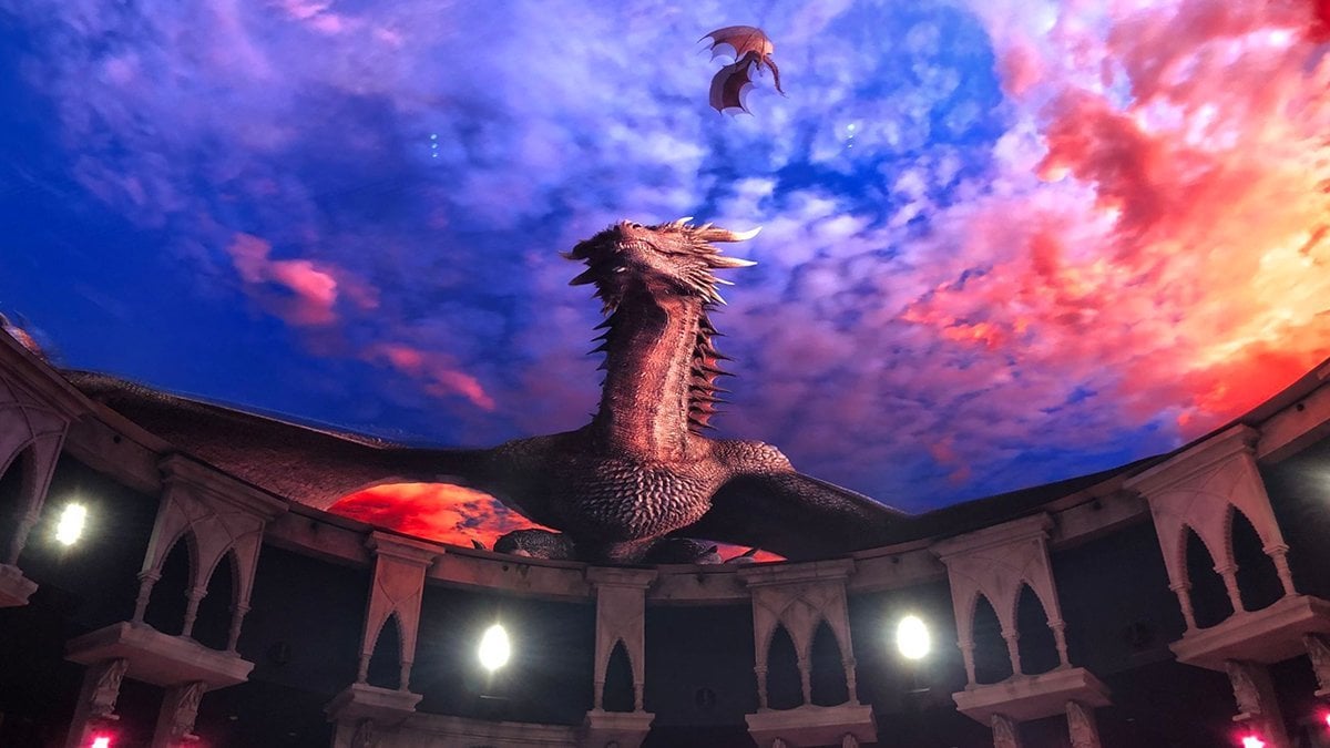 A large, detailed dragon emerges on an immersive, circular screen amidst a vibrant, fiery sky. A smaller dragon flies gracefully above. The setting echoes an arena with arches and lit torches—it's like stepping into a fantastical show at the Ron Clark Academy.