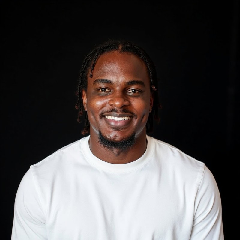 A smiling man with braided hair wearing a white shirt poses in front of a solid black background.