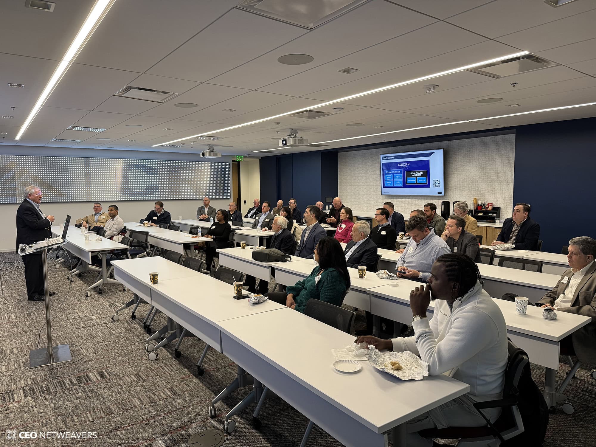 A business professional gives a presentation to a group of people seated at tables in a modern conference room. Attendees listen attentively, some with laptops, notebooks, and refreshments.