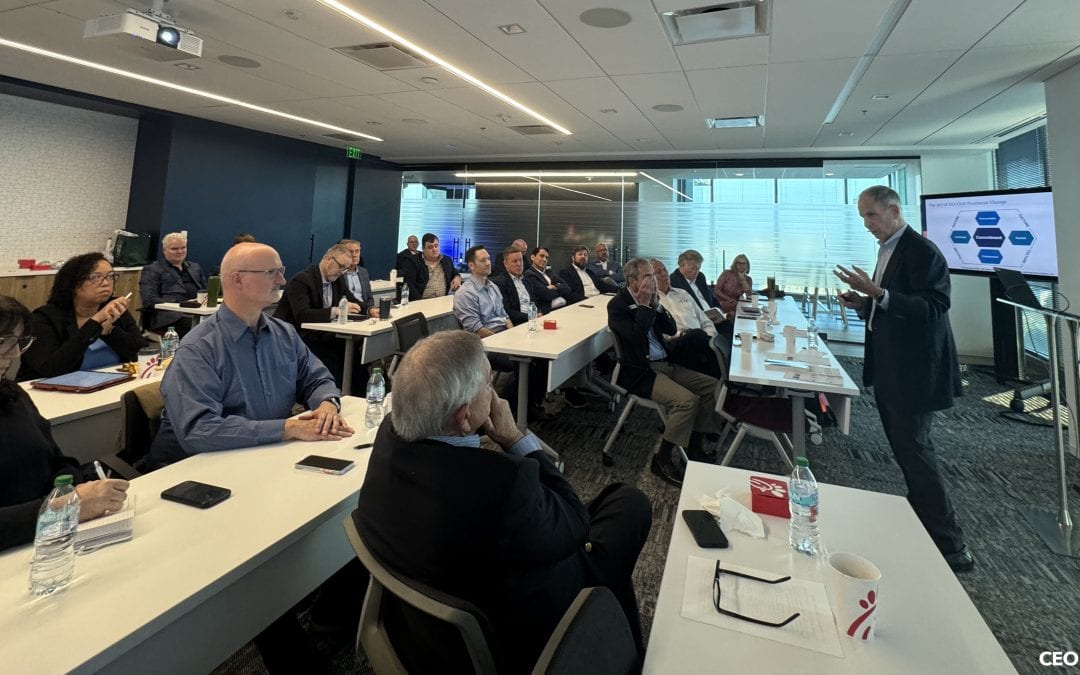 A group of professionals attends a business presentation in a modern conference room, listening to a man discuss healthy disagreement at the front near a screen displaying a diagram.