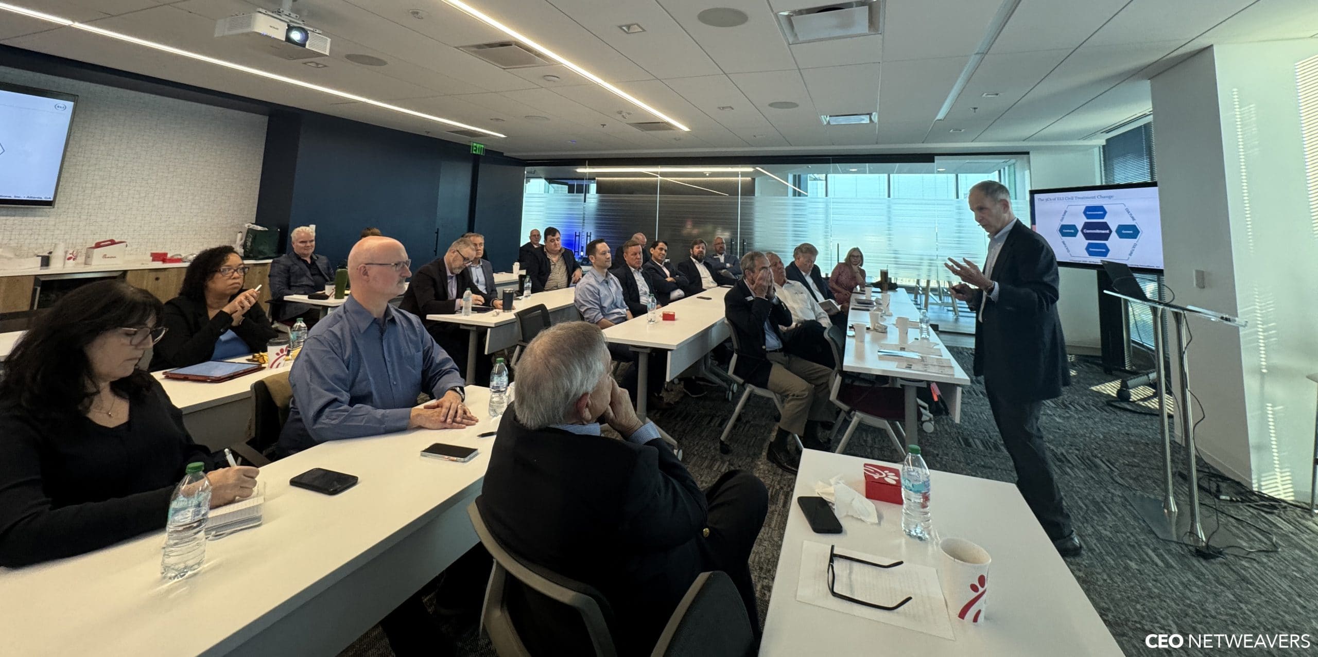 A group of professionals attends a business presentation in a modern conference room, listening to a man discuss healthy disagreement at the front near a screen displaying a diagram.